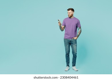 Full body european confident young man he wear purple t-shirt hold in hand use mobile cell phone surfint internet isolated on plain pastel light blue cyan background studio portrait. Lifestyle concept - Shutterstock ID 2308404461