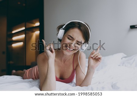 Full body excited caucasian young woman in pajamas headphones lying in bed listen to music dancing enjoy new playlist rest relax on weekends indoors at home. Good mood night morning bedtime concept.