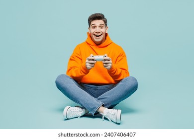 Full body excited fun young man wear orange hoody casual clothes sitting use play racing app on mobile cell phone hold gadget smartphone for pc video games isolated on plain blue cyan color background - Powered by Shutterstock