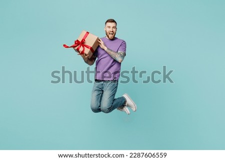 Full body excited fun young man he wears purple t-shirt jump high hold in hand present box with gift ribbon bow isolated on plain pastel light blue cyan background studio portrait. Lifestyle concept