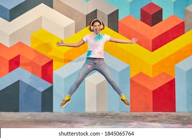 Full body of excited millennial female in casual outfit smeared with paint listening to music with headphones and jumping against wall with colorful geometric ornament - Shutterstock ID 1845065764