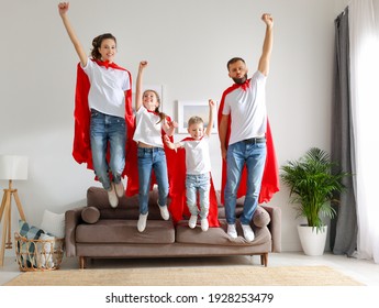 Full Body Excited Optimistic Family: Parents And Kids In Similar Casual Clothes And Red Superhero Cloaks Keeping Fists Up And Jumping Together From Sofa While Having Fun At Home