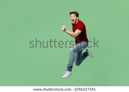 Full body excited overjoyed sporty young brunet man he wears red t-shirt casual clothes jump high run fast hurry up isolated on plain pastel light green background studio portrait. Lifestyle concept