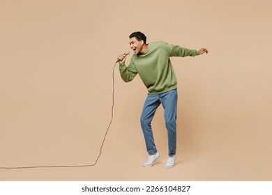 Full body expressive fun young man of African American ethnicity wear green sweatshirt sing song in microphone isolated on plain pastel light beige background studio portrait. People lifestyle concept - Shutterstock ID 2266104827