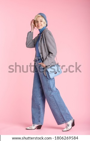 Full body fashion portrait of happy smiling woman wearing trendy wide leg jeans, houndstooth printed blazer, white cowboy boots, with light blue small shoulder bag, posing on pastel pink background

