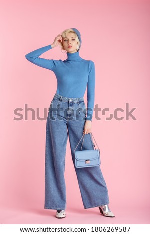 Full body fashion portrait of young elegant model wearing trendy wide leg jeans, light blue turtleneck, beret, holding small bag, posing on pastel pink background
