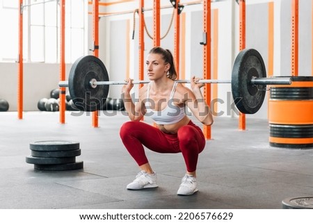 Full body female athlete in activewear looking away and doing barbell back squat during intense workout in sunlit gym