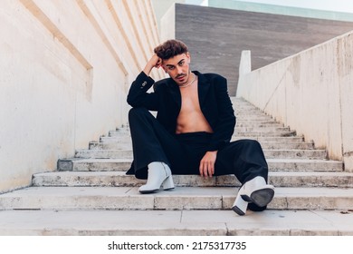 Full Body Of Feminine Male With Rhinestones On Face In High Heeled Boots Looking At Camera While Sitting On Staircase In City