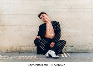 Full Body Of Feminine Male With Rhinestones On Face In High Heels Sitting Near Shabby Wall With Closed Eyes Under Sunlight In City