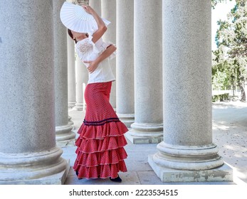 Full Body Flamenco Dancer With White Fan Covering Her Face. Dressed In A Red Skirt And Hugging Herself With One Arm.