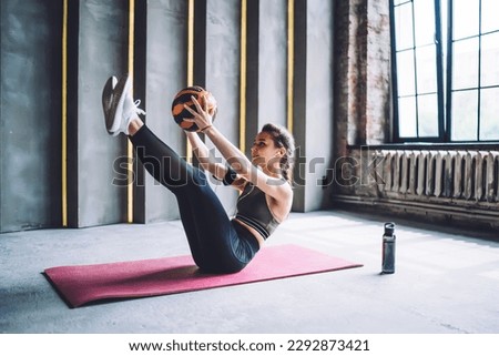 Full body of focused woman in sportswear sitting on mat and doing working out with ball while having abs workout in gym