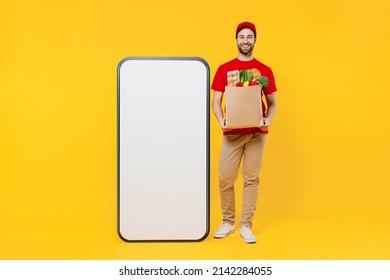 Full Body Fun Delivery Guy Employee Man Wear Red Cap T-shirt Uniform Work As Dealer Courier Stand Near Mobile Phone With Blank Screen Workspace Area Hold Food Bag Isolated On Plain Yellow Background