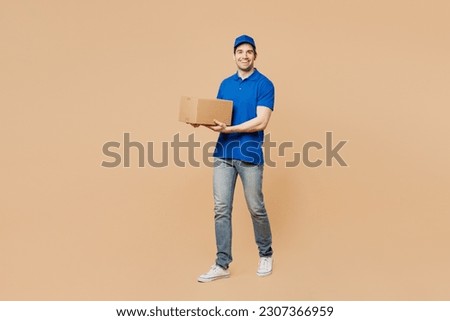 Full body fun delivery guy employee man wear blue cap t-shirt uniform workwear work as dealer courier hold cardboard box look camera go walk isolated on plain light beige background. Service concept