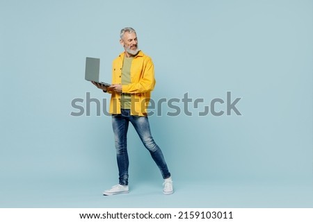 Full body fun elderly gray-haired mustache bearded man 50s wear yellow shirt hold use work on laptop pc computer look aside on workspace walk go isolated on plain pastel light blue background studio.