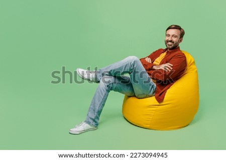 Full body fun elderly man 40s years old he wears casual clothes red shirt t-shirt sit in bag chair hold hands crossed folded look camera isolated on plain pastel light green background studio portrait