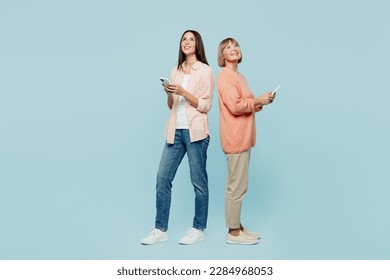 Full body fun happy elder parent mom with young adult daughter two women together wear casual clothes hold in hand mobile cell phone look overhead isolated on plain blue background. Family day concept