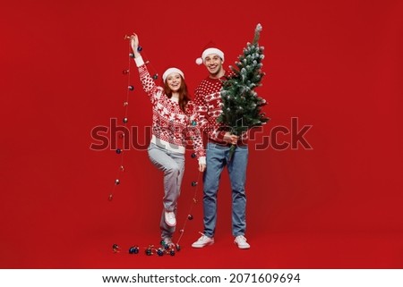 Full body fun happy young couple friends two man woman 20s in sweater hat hold fir-tree garland isolated on plain red background studio. Happy New Year 2022 celebration merry ho x-mas holiday concept