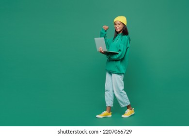 Full body fun IT little kid teen girl of African American ethnicity 13-14 years old wear casual hoody hat hold use work on laptop pc computer do winner gesture isolated on plain dark green background - Shutterstock ID 2207066729