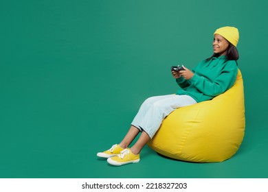 Full body fun little kid teen girl of African American ethnicity 13-14 years old wear casual hoody hat sit in bag chair hold play pc game with joystick console isolated on plain dark green background - Shutterstock ID 2218327203