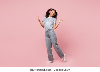 Full body fun little kid teen girl wears blue striped t-shirt casual clothes listen to music in headphones use mobile cell phone sing isolated on plain pastel light pink background. Childhood concept - Powered by Shutterstock