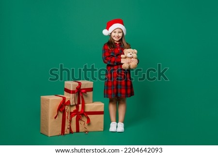 Full body fun merry little child kid girl 7 year old wear red dress Christmas hat posing near present box gift ribbon bow hold teddy bear isolated on plain green background Happy New Year 2023 concept
