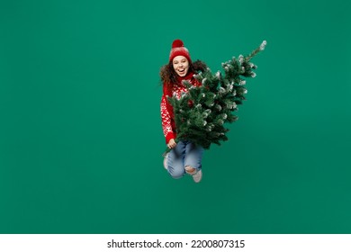 Full Body Fun Merry Young Woman Wearing Red Warm Cozy Knitted Sweater Hat Posing Jump High Hold Christmas Tree Isolated On Plain Dark Green Background. Happy New Year 2023 Celebration Holiday Concept