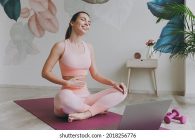 Full body fun satisfied happy smiling young sporty fitness trainer instructor woman in pink tracksuit do yoga exercise use laptop pc computer close eyes hold tummy sit on mat floor at home gym indoor - Shutterstock ID 2018256776