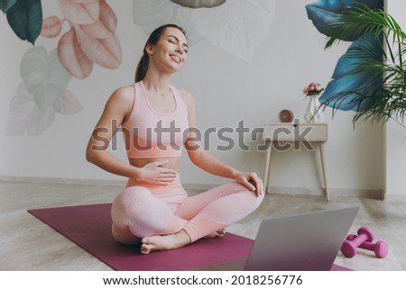 Full body fun satisfied happy smiling young sporty fitness trainer instructor woman in pink tracksuit do yoga exercise use laptop pc computer close eyes hold tummy sit on mat floor at home gym indoor