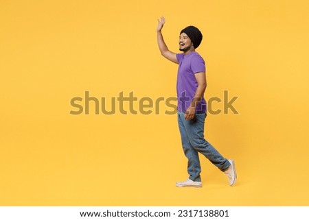 Full body fun side view smiling cheerful devotee Sikh Indian man ties his traditional turban dastar wear purple t-shirt walking going waving hand isolated on plain yellow background studio portrait