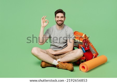 Full body fun smiling young traveler white man near backpack stuff mat sit show okay isolated on plain green background. Tourist leads active healthy lifestyle. Hiking trek rest travel trip concept