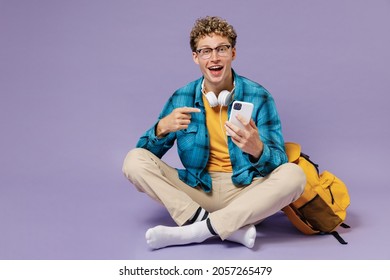 Full Body Fun Young Boy Teen Student Wear Casual Clothes Backpack Headphones Glasses Sit Point On Mobile Phone Isolated On Violet Background Studio Education In High School University College Concept.