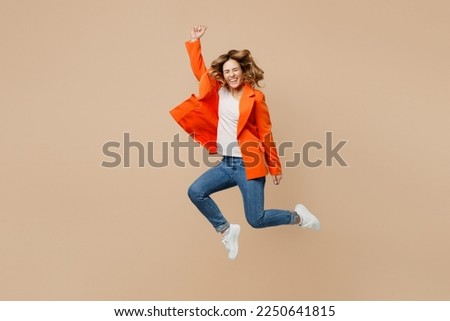 Full body fun young employee business woman corporate lawyer 30s wears classic formal orange suit glasses work in office jump high do winner gesture raise up hand isolated on plain beige background