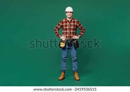 Full body fun young employee laborer handyman man wear red shirt hardhat stand akimbo hands on waist isolated on plain green background. Instruments for renovation apartment room. Repair home concept