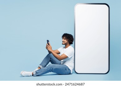 Full body fun young Indian man wear white t-shirt casual clothes sit near big huge blank screen mobile cell phone with area use smartphone isolated on plain pastel blue background. Lifestyle concept - Powered by Shutterstock