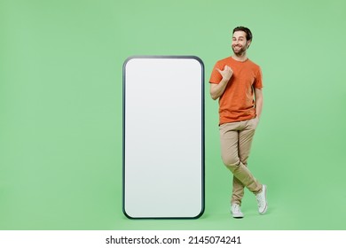 Full body fun young man 20s wear orange t-shirt stand point finger on mobile cell phone with blank screen workspace area isolated on plain pastel light green color background People lifestyle concept