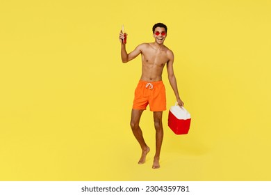 Full body fun young man wear shorts swimsuit glasses hold in hands beer bootle beach refrigerator relax near hotel pool isolated on plain yellow background. Summer vacation sea rest sun tan concept