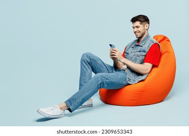 Full body fun young man he wear denim vest red t-shirt casual clothes sit in bag chair hold in hand use mobile cell phone isolated on plain pastel light blue cyan background studio. Lifestyle concept - Powered by Shutterstock