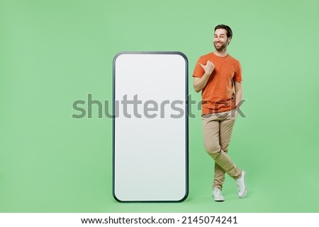 Full body fun young man 20s wear orange t-shirt stand point finger on mobile cell phone with blank screen workspace area isolated on plain pastel light green color background People lifestyle concept