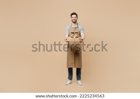 Full body fun young man barista barman employee wear brown apron work in coffee shop hold italian pizza in cardboard flatbox cup isolated on plain light beige background Small business startup concept