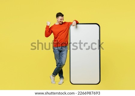Full body fun young man wear orange casual clothes stand near big huge blank screen mobile cell phone smartphone with copy space mockup area do winer gesture isolated on plain yellow color background