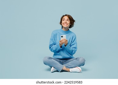 Full body fun young woman wear knitted sweater hold in hand use mobile cell phone look aside on workspace isolated on plain pastel light blue cyan background studio portrait. People lifestyle concept - Shutterstock ID 2242519583