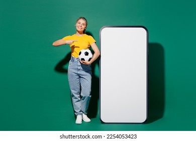 Full body fun young woman fan wears yellow t-shirt cheer up support football sport team hold soccer ball watch tv live stream show big blank screen mobile cell phone isolated on dark green background - Powered by Shutterstock