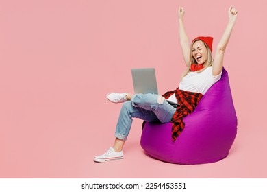 Full body fun young IT woman wear white t-shirt red hat sit in bag chair hold use work on laptop pc computer do winner gesture isolated on plain pastel light pink background. People lifestyle concept - Shutterstock ID 2254453551