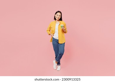Full body fun young woman of Asian ethnicity wear yellow shirt white t-shirt hold takeaway delivery craft paper brown cup coffee to go isolated on plain pastel light pink background studio portrait