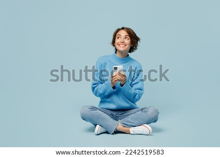 Photo of Full body fun young woman wear knitted sweater hold in hand use mobile cell phone look aside on workspace isolated on plain pastel light blue cyan background studio portrait. People lifestyle concept