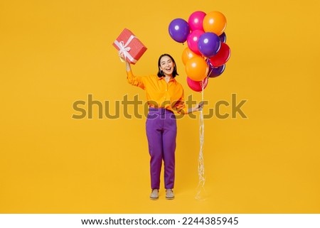 Full body fun young woman wear casual clothes hat celebrating hold bunch of balloons raise up present box with gift ribbon bow isolated on plain yellow background. Birthday 8 14 holiday party concept