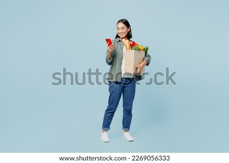 Full body fun young woman wear casual clothes hold brown paper bag with food products use mobile cell phone isolated on plain blue background studio portrait. Delivery service from shop or restaurant