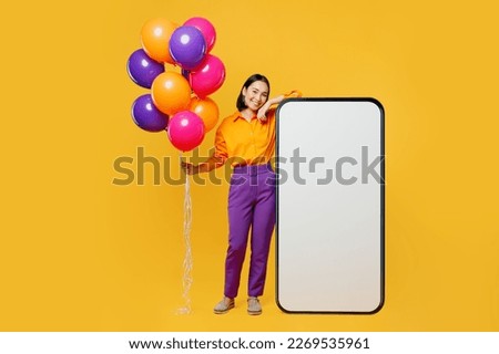 Full body fun young woman wear casual clothes hat celebrating with balloons near big huge blank screen area mobile cell phone isolated on plain yellow background. Birthday 8 14 holiday party concept