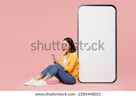 Full body fun young woman of Asian ethnicity wear yellow shirt white t-shirt sit near big huge blank screen mobile cell phone with area use smartphone isolated on plain pastel light pink background