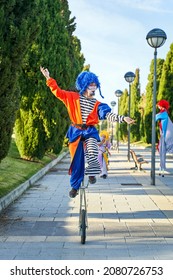 Full Body Of Funny Showman In Clown Costume And Blue Wig Riding Unicycle On Paved Pathway In Park While Performing Show With Anonymous Colleagues